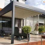 Zdjęcie prezentuje nowoczesną pergolę tarasową Luxury w zabudowie przyściennej, zamontowaną na przestronnym tarasie wyłożonym jasnymi płytami gresowymi. Aluminiowa konstrukcja w kolorze ciemnografitowym wspiera się na dwóch solidnych profilach frontowych, a jej dach stanowi jasnokremowe, segmentowe poszycie tkaninowe, które jest w pełni rozłożone. Pod zadaszeniem zaaranżowano strefę wypoczynkową z szarą sofą ogrodową oraz dwoma ciemnozielonymi fotelami. W tle widoczna jest jasna elewacja budynku z drewnianymi wstawkami, duże przeszklenia drzwi tarasowych oraz biała, dekoracyjna kratka z pnącymi roślinami. Na pierwszym planie znajdują się ozdobne drzewka na pniu, posadzone wzdłuż krawędzi tarasu wykończonej ciemnobrązową podmurówką z desek. Całość kompozycji w świetle dziennym podkreśla estetykę i funkcjonalność ochrony przeciwsłonecznej tarasu.