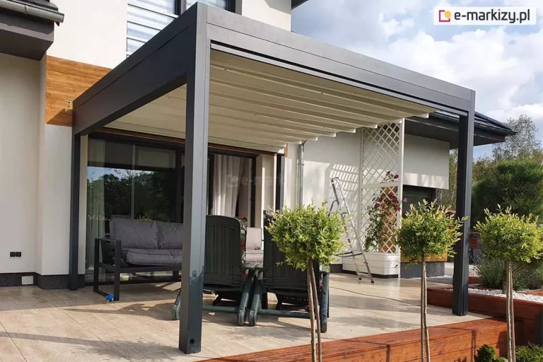 Zdjęcie prezentuje nowoczesną pergolę tarasową Luxury w zabudowie przyściennej, zamontowaną na przestronnym tarasie wyłożonym jasnymi płytami gresowymi. Aluminiowa konstrukcja w kolorze ciemnografitowym wspiera się na dwóch solidnych profilach frontowych, a jej dach stanowi jasnokremowe, segmentowe poszycie tkaninowe, które jest w pełni rozłożone. Pod zadaszeniem zaaranżowano strefę wypoczynkową z szarą sofą ogrodową oraz dwoma ciemnozielonymi fotelami. W tle widoczna jest jasna elewacja budynku z drewnianymi wstawkami, duże przeszklenia drzwi tarasowych oraz biała, dekoracyjna kratka z pnącymi roślinami. Na pierwszym planie znajdują się ozdobne drzewka na pniu, posadzone wzdłuż krawędzi tarasu wykończonej ciemnobrązową podmurówką z desek. Całość kompozycji w świetle dziennym podkreśla estetykę i funkcjonalność ochrony przeciwsłonecznej tarasu.