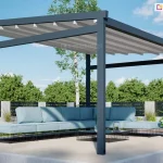 Wolnostojąca pergola ogrodowa Moderno o solidnej konstrukcji aluminiowej w kolorze ciemnego antracytu, wyposażona w rozsuwane zadaszenie tkaninowe w kolorze jasnoszarym. Materiał dachu układa się w charakterystyczne, estetyczne segmenty, wspierając się na poprzecznych profilach. Pod pergolą, na betonowym podeście, ustawiono nowoczesny narożnik ogrodowy z jasnoniebieskimi poduszkami na ażurowych, czarnych podstawach oraz minimalistyczny stolik kawowy. W tle widoczny jest zadbany ogród z zielonymi drzewami, trawnikiem oraz nowoczesnym ogrodzeniem panelowym, co podkreśla rekreacyjny charakter zabudowy.