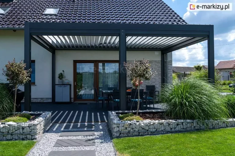 Nowoczesna, wolnostojąca pergola tarasowa Sb400 Aluprof o konstrukcji aluminiowej w kolorze antracytowym, usytuowana przy domu z dachem krytym czarną dachówką i jasną elewacją. Dach pergoli składa się z ruchomych lameli bioklimatycznych w kolorze białym, które są częściowo otwarte, rzucając pasiaste cienie na ciemny taras. Pod zadaszeniem widoczny jest stół z krzesłami oraz fragment elewacji wyłożonej jasnym kamieniem dekoracyjnym. Na pierwszym planie znajdują się gabiony wypełnione jasnym kamieniem, z których wyrastają ozdobne trawy oraz dwa młode drzewka liściaste. Przez środek ogrodu prowadzi ścieżka z szarych, prostokątnych płyt ułożonych na jasnym żwirze, otoczona zielonym trawnikiem. W tle widoczne są fragmenty sąsiednich zabudowań pod błękitnym niebem z białymi chmurami.