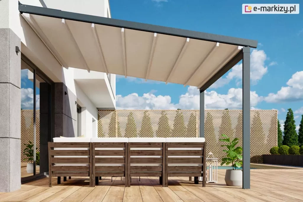 Nowoczesna pergola tarasowa Luxo na wymiar zamontowana do białej elewacji piętrowego budynku o minimalistycznej architekturze. Konstrukcja wsparta jest na dwóch pionowych, antracytowych słupach o kwadratowym przekroju, które stabilizują wysunięte zadaszenie nad przestronnym tarasem wykonanym z jasnych desek kompozytowych. Zadaszenie składa się z kremowej, lekko pofalowanej tkaniny rozpiętej na aluminiowych profilach poprzecznych, zapewniając ochronę przed słońcem nad strefą wypoczynkową. Pod pergolą ustawiono duży narożnik z ciemnego drewna z jasnymi poduchami, a obok znajduje się minimalistyczna donica z rośliną i dekoracyjna latarnia. W tle widoczne jest ażurowe ogrodzenie panelowe z jasnego drewna, rządek formowanych krzewów oraz błękitne niebo z rozproszonymi białymi chmurami, co podkreśla letni i wypoczynkowy charakter aranżacji.