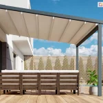 Nowoczesna pergola tarasowa Luxo na wymiar zamontowana do białej elewacji piętrowego budynku o minimalistycznej architekturze. Konstrukcja wsparta jest na dwóch pionowych, antracytowych słupach o kwadratowym przekroju, które stabilizują wysunięte zadaszenie nad przestronnym tarasem wykonanym z jasnych desek kompozytowych. Zadaszenie składa się z kremowej, lekko pofalowanej tkaniny rozpiętej na aluminiowych profilach poprzecznych, zapewniając ochronę przed słońcem nad strefą wypoczynkową. Pod pergolą ustawiono duży narożnik z ciemnego drewna z jasnymi poduchami, a obok znajduje się minimalistyczna donica z rośliną i dekoracyjna latarnia. W tle widoczne jest ażurowe ogrodzenie panelowe z jasnego drewna, rządek formowanych krzewów oraz błękitne niebo z rozproszonymi białymi chmurami, co podkreśla letni i wypoczynkowy charakter aranżacji.