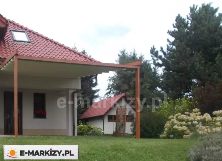 Nowoczesna pergola przyścienna z rozsuwanym dachem tkaninowym zamontowana na froncie domu jednorodzinnego o jasnej elewacji. Konstrukcja wykonana jest z profili aluminiowych w kolorze naturalnego drewna, co harmonizuje z dachem budynku pokrytym czerwoną dachówką ceramiczną. Zadaszenie z jasnej tkaniny wspiera się na dwóch pionowych słupach nośnych i jest częściowo wysunięte, tworząc osłonę nad tarasem przylegającym do bryły budynku. W tle widoczny jest zadbany, zielony ogród z gęstą roślinnością, drzewami iglastymi oraz mniejszym budynkiem gospodarczym o spójnej kolorystyce. Zdjęcie wykonane z perspektywy trawnika ukazuje integrację pergoli z otoczeniem oraz jej funkcję jako estetycznego przedłużenia strefy wypoczynkowej domu.