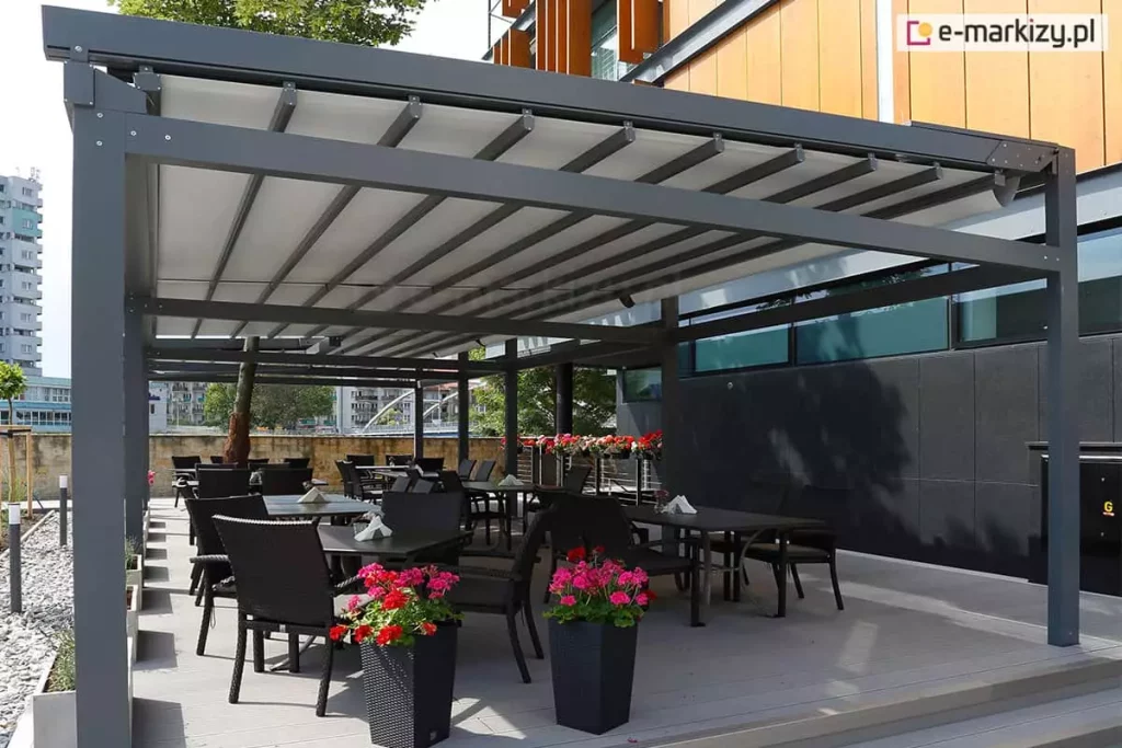 Nowoczesna, wielomodułowa pergola tarasowa o konstrukcji aluminiowej w kolorze antracytowym, stanowiąca zadaszenie ogródka restauracyjnego. Dach pergoli Solid Aluprof wykonany jest z jasnoszarego, segmentowego materiału rzymskiego, który jest rozsunięty na prowadnicach. Pod zadaszeniem, na jasnym podeście, ustawiono liczne czarne stoliki z dopasowanymi fotelami z technorattanu. Przestrzeń jest ozdobiona wysokimi, ciemnymi donicami z intensywnie różowymi i czerwonymi kwiatami. W tle widoczny jest nowoczesny budynek z elewacją z jasnego drewna i ciemnych paneli oraz fragmenty miejskiej zabudowy z wieżowcem w oddali. Po lewej stronie, na obrzeżu tarasu, znajduje się pas jasnego żwiru z niskimi nasadzeniami roślinnymi. Całość oświetlona jest jasnym, dziennym światłem.