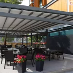 Nowoczesna, wielomodułowa pergola tarasowa o konstrukcji aluminiowej w kolorze antracytowym, stanowiąca zadaszenie ogródka restauracyjnego. Dach pergoli Solid Aluprof wykonany jest z jasnoszarego, segmentowego materiału rzymskiego, który jest rozsunięty na prowadnicach. Pod zadaszeniem, na jasnym podeście, ustawiono liczne czarne stoliki z dopasowanymi fotelami z technorattanu. Przestrzeń jest ozdobiona wysokimi, ciemnymi donicami z intensywnie różowymi i czerwonymi kwiatami. W tle widoczny jest nowoczesny budynek z elewacją z jasnego drewna i ciemnych paneli oraz fragmenty miejskiej zabudowy z wieżowcem w oddali. Po lewej stronie, na obrzeżu tarasu, znajduje się pas jasnego żwiru z niskimi nasadzeniami roślinnymi. Całość oświetlona jest jasnym, dziennym światłem.