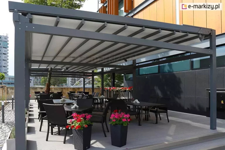 Nowoczesna, wielomodułowa pergola tarasowa o konstrukcji aluminiowej w kolorze antracytowym, stanowiąca zadaszenie ogródka restauracyjnego. Dach pergoli Solid Aluprof wykonany jest z jasnoszarego, segmentowego materiału rzymskiego, który jest rozsunięty na prowadnicach. Pod zadaszeniem, na jasnym podeście, ustawiono liczne czarne stoliki z dopasowanymi fotelami z technorattanu. Przestrzeń jest ozdobiona wysokimi, ciemnymi donicami z intensywnie różowymi i czerwonymi kwiatami. W tle widoczny jest nowoczesny budynek z elewacją z jasnego drewna i ciemnych paneli oraz fragmenty miejskiej zabudowy z wieżowcem w oddali. Po lewej stronie, na obrzeżu tarasu, znajduje się pas jasnego żwiru z niskimi nasadzeniami roślinnymi. Całość oświetlona jest jasnym, dziennym światłem.