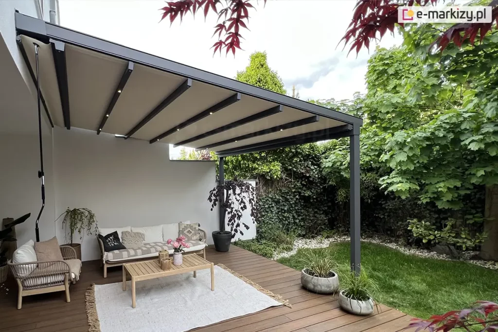 Zdjęcie prezentuje nowoczesną pergolę tarasową aura o konstrukcji aluminiowej w kolorze antracytowym, zamontowaną do jasnej elewacji budynku mieszkalnego. Pergola posiada rozsuwane zadaszenie tkaninowe w kolorze kremowym, które jest rozpięte na systemie poprzecznych profili jezdnych. W dolnej części profili zamontowano zintegrowane, punktowe oświetlenie LED, emitujące ciepłe światło. Konstrukcja wspiera się na pionowych słupach o kwadratowym przekroju, usytuowanych na rogu drewnianego tarasu. Pod pergolą zaaranżowano strefę wypoczynkową z meblami ogrodowymi o drewnianej strukturze i jasnych poduszkach, składającą się z sofy i fotela. Na meblach leżą poduszki dekoracyjne, w tym jedna czarna z białym napisem oraz jedna beżowa z etnicznym wzorem. Na tarasie z ciemnobrązowych desek leży jasny dywan z frędzlami, na którym stoi prostokątny, drewniany stolik kawowy z wazonem z różowymi kwiatami i dekoracyjnymi świecznikami. Po lewej stronie, przy ścianie budynku, zamocowana jest długa, czarna korba do manualnego sterowania zadaszeniem. W narożniku tarasu rośnie ozdobne drzewko o bordowych liściach w ciemnej donicy, a przy krawędzi ustawiono dwie okrągłe, betonowe donice z zielonymi trawami. Tło stanowi gęsty, zielony ogród z krzewami i drzewami liściastymi, a u góry kadru widoczne są gałązki z czerwonymi liśćmi, prawdopodobnie klonu palmowego, obramowujące ujęcie.