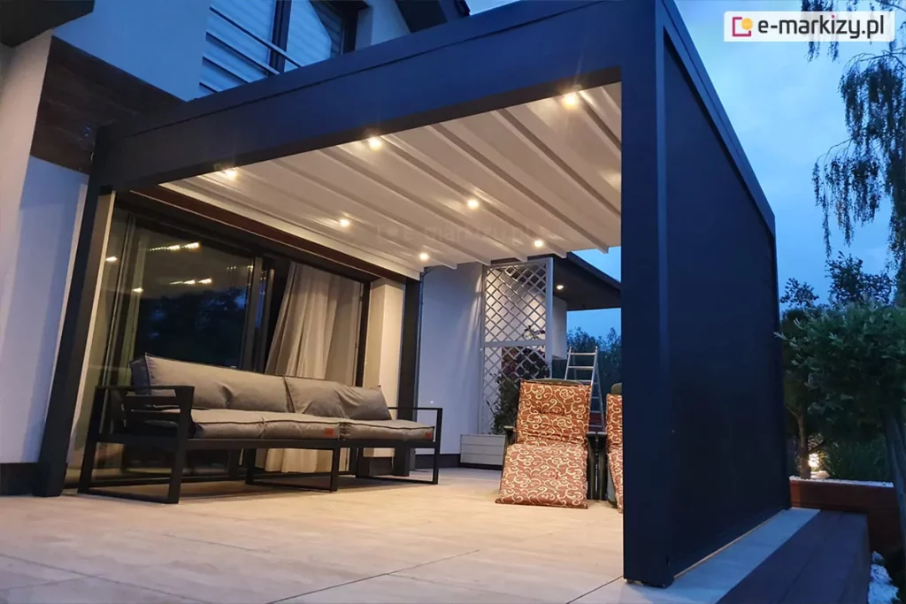 Zdjęcie prezentuje pergolę tarasową Luxury w wersji przyściennej, uchwyconą o zmierzchu z włączonym zintegrowanym oświetleniem punktowym LED. Konstrukcja pergoli wykonana jest z grubych profili aluminiowych w kolorze ciemnografitowym, a jej dach stanowi jasne, segmentowe poszycie materiałowe, które jest w pełni rozłożone. W poprzecznych belkach zadaszenia zamontowano szereg punktów świetlnych emitujących ciepłe światło, które oświetla przestrzeń wypoczynkową pod konstrukcją. Prawa strona pergoli została wyposażona w pełną ściankę boczną, prawdopodobnie w formie rolety typu screen, która domyka przestrzeń tarasu. Na jasnej powierzchni tarasu, wykonanej z dużych płyt, ustawiono szarą, nowoczesną sofę ogrodową na metalowym stelażu oraz fotele z wzorzystymi pomarańczowo-brązowymi poduszkami. W tle widoczna jest elewacja budynku z dużym przeszkleniem drzwi tarasowych, za którymi widać zasłony, oraz biała ozdobna kratka ogrodowa. Całość kompozycji podkreśla funkcjonalność oświetlenia dachowego w warunkach wieczornych.