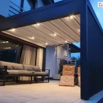 Zdjęcie prezentuje pergolę tarasową Luxury w wersji przyściennej, uchwyconą o zmierzchu z włączonym zintegrowanym oświetleniem punktowym LED. Konstrukcja pergoli wykonana jest z grubych profili aluminiowych w kolorze ciemnografitowym, a jej dach stanowi jasne, segmentowe poszycie materiałowe, które jest w pełni rozłożone. W poprzecznych belkach zadaszenia zamontowano szereg punktów świetlnych emitujących ciepłe światło, które oświetla przestrzeń wypoczynkową pod konstrukcją. Prawa strona pergoli została wyposażona w pełną ściankę boczną, prawdopodobnie w formie rolety typu screen, która domyka przestrzeń tarasu. Na jasnej powierzchni tarasu, wykonanej z dużych płyt, ustawiono szarą, nowoczesną sofę ogrodową na metalowym stelażu oraz fotele z wzorzystymi pomarańczowo-brązowymi poduszkami. W tle widoczna jest elewacja budynku z dużym przeszkleniem drzwi tarasowych, za którymi widać zasłony, oraz biała ozdobna kratka ogrodowa. Całość kompozycji podkreśla funkcjonalność oświetlenia dachowego w warunkach wieczornych.