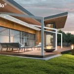Biała pergola przyścienna Tarasola Light z całkowicie zwiniętym zadaszeniem tkaninowym, zamontowana na nowoczesnym tarasie wyłożonym deską kompozytową. Widoczna jest lekka, aluminiowa konstrukcja szkieletowa składająca się z ukośnych, poziomych prowadnic oraz pionowych podpór, która integruje się z bryłą budynku o elewacji z płyt architektonicznych w kolorze miedzi i antracytu. W strefie wypoczynkowej pod pergolą ustawiono minimalistyczne meble tarasowe z szarymi siedziskami, a na pierwszym planie płonie ogień w czarnym, misowatym palenisku ogrodowym. Wieczorna sceneria z oświetloną trawą i lasem w tle ukazuje funkcjonalność oświetlenia punktowego LED zamontowanego w profilach pergoli, mimo że materiał dachu pozostaje ukryty pod daszkiem ochronnym. Całość aranżacji podkreśla luksusowy charakter przydomowej przestrzeni rekreacyjnej z widokiem na zachodzące, różowo-fioletowe niebo.
