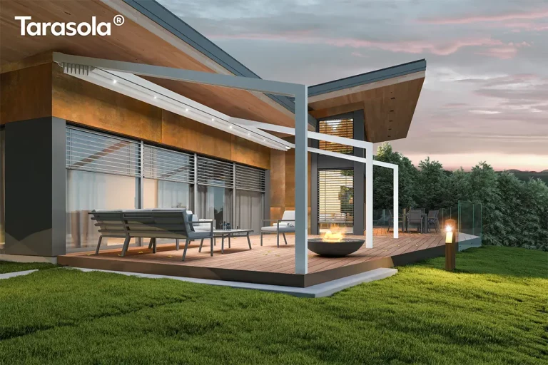 Biała pergola przyścienna Tarasola Light z całkowicie zwiniętym zadaszeniem tkaninowym, zamontowana na nowoczesnym tarasie wyłożonym deską kompozytową. Widoczna jest lekka, aluminiowa konstrukcja szkieletowa składająca się z ukośnych, poziomych prowadnic oraz pionowych podpór, która integruje się z bryłą budynku o elewacji z płyt architektonicznych w kolorze miedzi i antracytu. W strefie wypoczynkowej pod pergolą ustawiono minimalistyczne meble tarasowe z szarymi siedziskami, a na pierwszym planie płonie ogień w czarnym, misowatym palenisku ogrodowym. Wieczorna sceneria z oświetloną trawą i lasem w tle ukazuje funkcjonalność oświetlenia punktowego LED zamontowanego w profilach pergoli, mimo że materiał dachu pozostaje ukryty pod daszkiem ochronnym. Całość aranżacji podkreśla luksusowy charakter przydomowej przestrzeni rekreacyjnej z widokiem na zachodzące, różowo-fioletowe niebo.