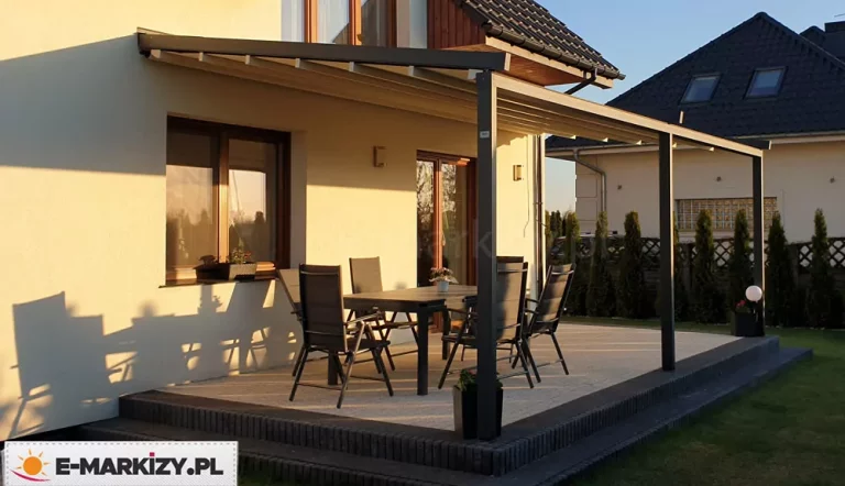 Nowoczesna pergola przyścienna Luxo z rozsuwanym zadaszeniem tkaninowym zamontowana na tarasie domku jednorodzinnego. Aluminiowa konstrukcja w ciemnym, antracytowym kolorze składa się z prowadnic przymocowanych do elewacji budynku oraz trzech pionowych słupów nośnych posadowionych na podwyższonym tarasie wyłożonym jasną kostką brukową. Zadaszenie wykonane z jasnej, kremowej tkaniny jest rozwinięte, ukazując system poprzecznych profili wzmacniających. Pod pergolą znajduje się zestaw mebli ogrodowych składający się z dużego stołu oraz sześciu krzeseł z wysokimi oparciami. Całość aranżacji dopełniają detale takie jak donice z roślinami oraz oświetlenie elewacyjne. Zdjęcie wykonane w świetle popołudniowego słońca, które rzuca wyraźne cienie konstrukcji na jasną ścianę budynku i nawierzchnię tarasu, podkreślając teksturę materiałów i stabilność budowli.