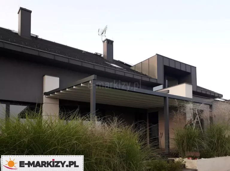 Nowoczesna pergola przyścienna Moderno o konstrukcji w kolorze ciemnego antracytu, zamontowana przy elewacji piętrowego budynku o grafitowych i beżowych ścianach. Zadaszenie tkaninowe w jasnym odcieniu jest w pełni rozwinięte, tworząc płaską płaszczyznę chroniącą taras. Konstrukcja zintegrowana jest z nowoczesną bryłą domu, charakteryzującą się ciemną dachówką i dużymi przeszkleniami. Na pierwszym planie widoczne są wysokie, ozdobne trawy ogrodowe, które częściowo przesłaniają dolną partię tarasu, nadając aranżacji naturalny i prywatny charakter.