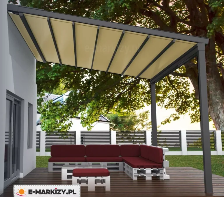 Nowoczesna pergola tarasowa o antracytowej, aluminiowej konstrukcji, zamontowana przy białej elewacji budynku. Zadaszenie pergoli składa się z jasnego, kremowego poszycia materiałowego, które jest rozpięte na systemie równoległych profili poprzecznych w kolorze ciemnoszarym. Konstrukcja wspiera się na dwóch solidnych słupach pionowych o przekroju kwadratowym. Pod pergolą, na tarasie z ciemnych, brązowych desek, znajduje się narożny zestaw mebli ogrodowych wykonany z białych palet z grubymi siedziskami i oparciami w kolorze bordowym. Przed narożnikiem ustawiono niski stolik oraz puf, również wykonane z pomalowanych na biało palet. W tle widoczne jest wysokie, nowoczesne ogrodzenie z ciemnych, poziomych lameli z białymi słupkami oraz duże, rozłożyste drzewo liściaste, którego gałęzie częściowo nachodzą nad zadaszenie. Scena ukazuje strefę wypoczynkową w świetle dziennym, z widocznym fragmentem białego budynku z szarą stolarką okienną po lewej stronie