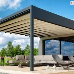 Zdjęcie prezentuje wizualizację luksusowej pergoli tarasowej Luxury o nowoczesnej, minimalistycznej konstrukcji aluminiowej w kolorze antracytowym. Zadaszenie stanowi dach tkaninowy w odcieniu jasnego beżu, który jest rozsunięty, tworząc miękkie, horyzontalne fale materiału rozpięte na prowadnicach. Konstrukcja wspiera się na solidnych profilach o kwadratowym przekroju i jest zamontowana do białej elewacji budynku z fragmentem wykonanym z czerwonej cegły. Pod pergolą, na jasnym drewnianym tarasie, zaaranżowano strefę wypoczynkową z dużym, niskim narożnikiem w kolorze taupe oraz dwoma designerskimi, ażurowymi fotelami w ciemnej tonacji. W przeszkleniach budynku odbija się błękitne niebo z białymi chmurami, a otoczenie tarasu wypełnia starannie wypielęgnowany ogród z ozdobnymi trawami i drzewami. Całość aranżacji uzupełniają stylowe detale, takie jak nowoczesne donice z roślinami oraz białe lampiony dekoracyjne ustawione na deskach tarasowych.