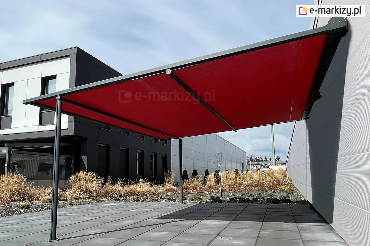 Lekka pergola tarasowa przyścienna o nowoczesnej konstrukcji aluminiowej w kolorze czarnym, zamontowana do elewacji budynku przemysłowego lub biurowego wykonanego z jasnoszarych paneli. Zadaszenie stanowi intensywnie czerwone, gładkie poszycie tkaninowe, które jest całkowicie rozpięte na bocznych prowadnicach i wsparte na poprzecznej belce frontowej. Konstrukcja opiera się na dwóch smukłych, pionowych słupach o okrągłym przekroju, przytwierdzonych do podłoża z dużych, ciemnoszarych płyt betonowych. W tle widoczna jest bryła nowoczesnego, czarnego budynku z regularnym układem okien oraz fragmenty niskiej roślinności i ozdobnych traw posadzonych wzdłuż elewacji. Zdjęcie wykonane w świetle dziennym przy lekko zachmurzonym niebie, prezentuje kontrastowe zestawienie kolorystyczne czerwonej tkaniny z surową, nowoczesną architekturą otoczenia.