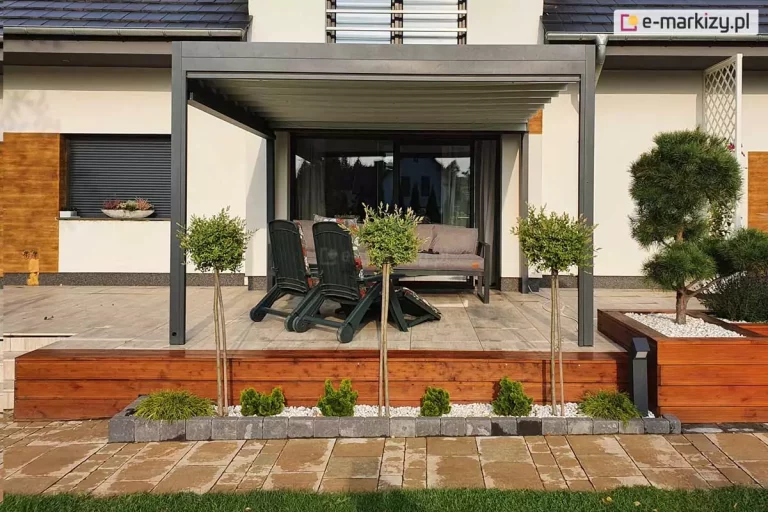Zdjęcie przedstawia nowoczesną pergolę tarasową Luxury o konstrukcji przyściennej, zamontowaną na podwyższonym tarasie wykonanym z jasnych płyt ceramicznych. Konstrukcja pergoli składa się z grafitowych, aluminiowych profili o matowym wykończeniu, z dwiema pionowymi nogami wspierającymi przednią część zadaszenia. Dach pergoli stanowi rozsuwane poszycie w kolorze jasnoszarym, które w tej chwili jest rozwinięte, ukazując segmentową budowę materiału wspartą na poprzecznych belkach. Taras jest osadzony na drewnianej podmurówce z ciemnobrązowych desek, przed którą znajduje się pas niskiej roślinności ogrodowej oraz ozdobne białe kamienie otoczone szarą kostką brukową. W strefie wypoczynkowej pod pergolą umieszczono szarą sofę oraz dwa ciemne, rozkładane fotele ogrodowe. W tle widoczna jest elewacja nowoczesnego domu z elementami drewna, dużymi przeszkleniami oraz zewnętrzną roletą okienną. Po prawej stronie tarasu znajduje się drewniana donica z formowanym drzewkiem typu bonsai oraz dekoracyjne białe kratki przy ścianie budynku.