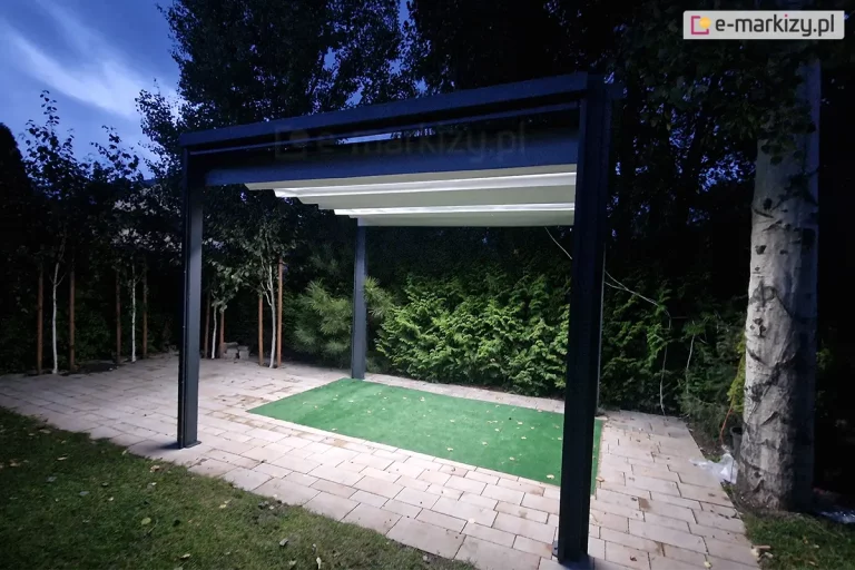 Nowoczesna, wolnostojąca pergola ogrodowa w wersji zintegrowanej z oświetleniem LED, sfotografowana wieczorem. Antracytowa konstrukcja aluminiowa wyposażona jest w jasne, kremowe zadaszenie rzymskie, pod którym zamontowano liniowe źródła światła, oświetlające przestrzeń pod pergolą. Obiekt ustawiony jest na utwardzonym podłożu z jasnej kostki brukowej, z centralnie umieszczonym pasem zielonej sztucznej trawy. Wokół pergoli znajduje się gęsta roślinność ogrodowa, w tym tuje oraz drzewa liściaste o jasnej korze, które są delikatnie doświetlone blaskiem wydobywającym się spod zadaszenia. Kontrast między ciemnym otoczeniem a jasnym, ciepłym światłem wewnątrz konstrukcji podkreśla ekskluzywny charakter strefy relaksu. Zdjęcie prezentuje funkcjonalność pergoli jako całorocznego elementu architektury ogrodowej, zdatnego do użytku również po zachodzie słońca.