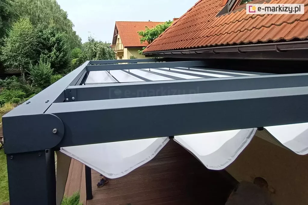 Pergola rzymska Tako przymocowana do ściany budynku tuz pod spadzistym dachem pokrytym czerwoną dachówką. Aluminiowa konstrukcja pomalowana na kolor antracytowy kontrastuje z białą tkaniną. Pd pergolą widoczny fragment tarasu wyłożonego drewnianymi deskami, po prawej odwrócony do ściany mężczyzna, pod dachem widoczne dwie stopy w butach sportowych. Ponad pergolą widoczne liczne zielone drzewa oraz fragment sąsiedniego budynku.