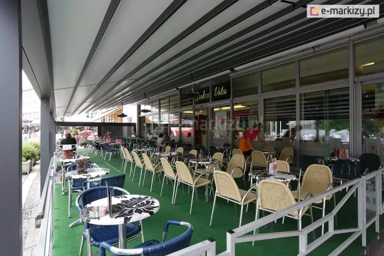 Zdjęcie prezentuje zastosowanie pergoli rzymskiej model Solid Aluprof jako zadaszenia ogródka restauracyjnego "Dolce Vita". Konstrukcja o imponującej rozpiętości wykonana jest z profili aluminiowych w kolorze antracytowym i została zamontowana wzdłuż przeszklonej fasady lokalu. Zadaszenie stanowi jasna, segmentowa tkanina, która jest całkowicie rozłożona, chroniąc dużą powierzchnię stolików przed słońcem lub deszczem. Przestrzeń pod pergolą jest wyposażona w liczne zestawy kawiarniane: stoliki z blatami w biało-czarne wzory kwiatowe oraz krzesła w kolorach beżowym i granatowym. Podłoże wyłożone jest estetyczną, zieloną wykładziną imitującą trawę. Ogródek jest oddzielony od chodnika niską, białą barierką o geometrycznym wzorze. Wewnątrz lokalu, za dużymi witrynami, widoczna jest obsługa oraz nowoczesne oświetlenie wnętrza. Perspektywa zdjęcia ukazuje rytmiczny układ profili dachu, który optycznie wydłuża przestrzeń restauracyjną.