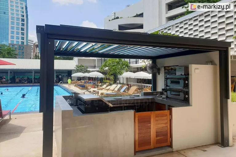 Wolnostojąca pergola bioklimatyczna SB400 w kolorze antracytowym, zainstalowana jako zadaszenie zewnętrznej kuchni letniej przy basenie. Konstrukcja posiada dach z ruchomych lameli ustawionych pod kątem, co zapewnia wentylację i półcień. Pod pergolą znajduje się stanowisko gastronomiczne z betonową zabudową, w której osadzono dwupoziomowy piec do pizzy, blaty robocze oraz szafki. Po pergolą przestrzeń baru ograniczona została betonowymi, niskimi ściankami, z drewnianymi drzwiami uchylnymi. W tle widoczny jest duży basen rekreacyjny z wyznaczonymi torami do pływania, otoczony strefą wypoczynku z licznymi leżakami i białymi parasolami. Całość usytuowana jest w sąsiedztwie nowoczesnej infrastruktury hotelowej oraz wysokich budynków miejskich pod czystym niebem.