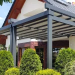 Zdjęcie prezentuje szeroką pergolę tarasową model Solid Aluprof o konstrukcji aluminiowej w kolorze ciemnego antracytu, zamontowaną do elewacji dużego domu jednorodzinnego. Zadaszenie składa się z dwóch niezależnych modułów tkaninowych w kolorze jasnokremowym, które są rozpięte na systemie poprzecznych profili aluminiowych. Konstrukcja wspiera się na trzech masywnych słupach frontowych o przekroju kwadratowym. Budynek posiada jasną elewację, fragmenty wykończone czerwoną cegłą klinkierową oraz ciemną dachówkę. Pergola osłania przestronny taras, a w jej bezpośrednim sąsiedztwie znajdują się gęste, formowane krzewy ozdobne (tuje) oraz inna niska roślinność ogrodowa. Całość kompozycji ukazana jest w pełnym świetle dziennym na tle bezchmurnego nieba, co podkreśla nowoczesny charakter instalacji i jej integrację z architekturą domu.