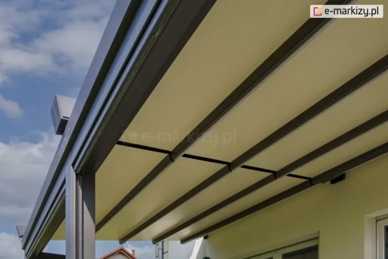 zbliżenie na dolną część zadaszenia pergoli model Solid Aluprof w widoku od strony tarasu. Konstrukcja zadaszenia opiera się na równolegle rozmieszczonych profilach poprzecznych w kolorze ciemnego antracytu, które podtrzymują gładko napiętą tkaninę w kolorze jasnokremowym. Materiał jest całkowicie rozłożony, tworząc jednolitą i płaską powierzchnię chroniącą przed słońcem. Po lewej stronie widoczny jest masywny profil boczny z prowadnicą, w której poruszają się elementy dachu. W górnym lewym rogu widać fragment zakończenia szyny jezdnej o geometrycznym kształcie. Tło stanowi jasna, tynkowana elewacja budynku z fragmentem ramy okiennej oraz błękitne niebo z rozproszonymi, białymi chmurami. Perspektywa zdjęcia z dołu podkreśla rytmiczny układ profili konstrukcyjnych i precyzyjne napięcie poszycia.
