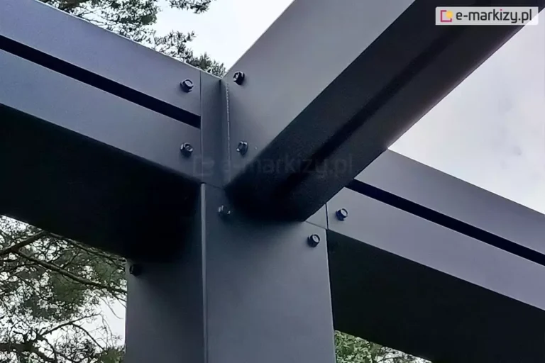 Fragment konstrukcji pergoli Tako, łączenie słupa pergoli z krokwią dachową za pomocą trójnika. Łebki śrub wystają poza elementy konstrukcji w kolorze antracytowym. W tle widoczne gałęzie drzew i niebo.