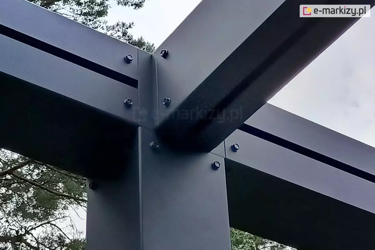 Fragment konstrukcji pergoli Tako, łączenie słupa pergoli z krokwią dachową za pomocą trójnika. Łebki śrub wystają poza elementy konstrukcji w kolorze antracytowym. W tle widoczne gałęzie drzew i niebo.