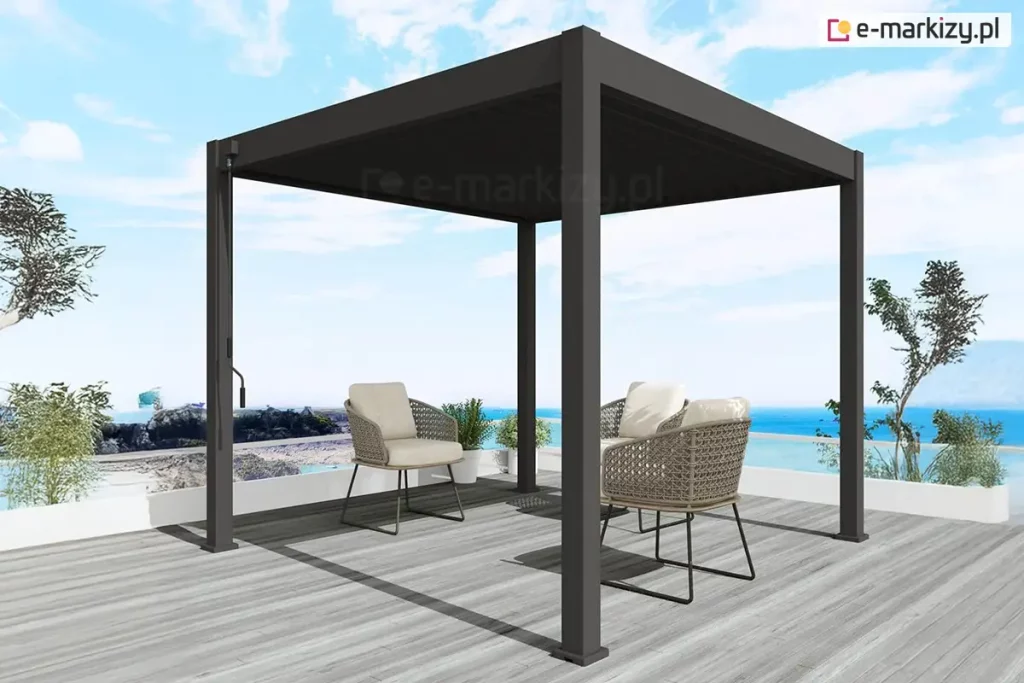 Pergola aluminiowa z dachem lamelowym Marlo w kolorze antracytowym, z napędem ręcznym korbowym, ustawiona na tarasie z jasnoszarych desek z widokiem na morze, skały i drzewa. Pod pergolą w pobliżu niskiej, szklanej barierki tarasu ustawiono trzy krzesła z plecionym oparciem, poduszkami na cienkich metalowych podporach.