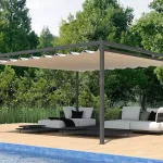 Wizualizacja wolnostojącej pergoli ogrodowej o minimalistycznej, antracytowej konstrukcji aluminiowej, ustawionej na jasnym, drewnianym tarasie przy basenie. Pergola tarasowa Dragon wolnostojąca posiada dach wykonany z jasnobeżowej tkaniny akrylowej, który jest rozłożony, tworząc regularne, dekoracyjne pofalowania między poprzecznymi profilami prowadzącymi. Pod pergolą zaaranżowano nowoczesną strefę wypoczynkową z białymi, niskimi modułowymi sofami ozdobionymi ciemnymi poduszkami. Meble spoczywają na płaskich, czarnych platformach, które pełnią funkcję zintegrowanych stolików bocznych. W dolnej części kadru widoczny jest fragment basenu z błękitną mozaiką oraz dwa brązowe, ergonomiczne leżaki ustawione na brzegu tarasu. Tło stanowi gęsta ściana wysokich, zielonych drzew oraz pas niskich traw ozdobnych, które oddzielają strefę tarasu od naturalnego krajobrazu. Całość ujęcia prezentuje się w pełnym, dziennym świetle, podkreślając luksusowy charakter letniej strefy relaksu.