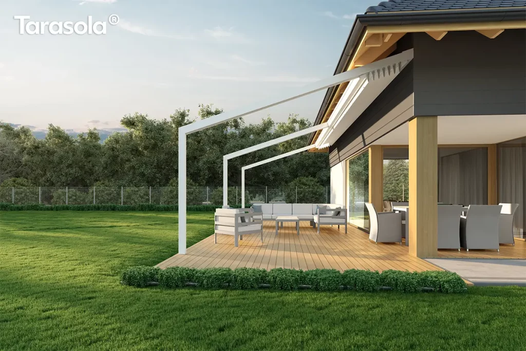 Biała pergola przyścienna Tarasola Light w pełnym świetle dziennym, zamontowana nad jasnym drewnianym tarasem przylegającym do nowoczesnego domu o grafitowej elewacji z drewnianymi filarami. Aluminiowa konstrukcja o pochyłym dachu posiada całkowicie zwinięte zadaszenie tkaninowe, które w tej pozycji tworzy dekoracyjne plisy pod okapem budynku. Na tarasie zaaranżowano przestronną strefę wypoczynkową z dużą narożną sofą w kolorze jasnoszarym, niskim stolikiem kawowym oraz kompletem jadalnianym z fotelami o technorattanowej strukturze ukrytymi w podcieniu. Teren wokół tarasu wypełnia zadbany trawnik, niska roślinność ozdobna oddzielająca deski od trawy oraz gęsty pas zielonych drzew w tle, a cała scena skąpana jest w miękkim słońcu przy bezchmurnym, błękitnym niebie.