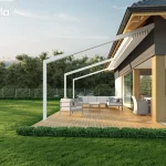 Biała pergola przyścienna Tarasola Light w pełnym świetle dziennym, zamontowana nad jasnym drewnianym tarasem przylegającym do nowoczesnego domu o grafitowej elewacji z drewnianymi filarami. Aluminiowa konstrukcja o pochyłym dachu posiada całkowicie zwinięte zadaszenie tkaninowe, które w tej pozycji tworzy dekoracyjne plisy pod okapem budynku. Na tarasie zaaranżowano przestronną strefę wypoczynkową z dużą narożną sofą w kolorze jasnoszarym, niskim stolikiem kawowym oraz kompletem jadalnianym z fotelami o technorattanowej strukturze ukrytymi w podcieniu. Teren wokół tarasu wypełnia zadbany trawnik, niska roślinność ozdobna oddzielająca deski od trawy oraz gęsty pas zielonych drzew w tle, a cała scena skąpana jest w miękkim słońcu przy bezchmurnym, błękitnym niebie.