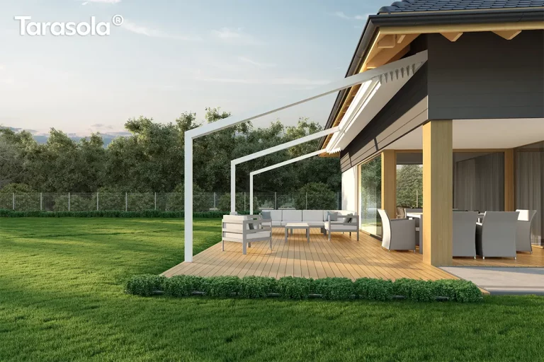 Biała pergola przyścienna Tarasola Light w pełnym świetle dziennym, zamontowana nad jasnym drewnianym tarasem przylegającym do nowoczesnego domu o grafitowej elewacji z drewnianymi filarami. Aluminiowa konstrukcja o pochyłym dachu posiada całkowicie zwinięte zadaszenie tkaninowe, które w tej pozycji tworzy dekoracyjne plisy pod okapem budynku. Na tarasie zaaranżowano przestronną strefę wypoczynkową z dużą narożną sofą w kolorze jasnoszarym, niskim stolikiem kawowym oraz kompletem jadalnianym z fotelami o technorattanowej strukturze ukrytymi w podcieniu. Teren wokół tarasu wypełnia zadbany trawnik, niska roślinność ozdobna oddzielająca deski od trawy oraz gęsty pas zielonych drzew w tle, a cała scena skąpana jest w miękkim słońcu przy bezchmurnym, błękitnym niebie.