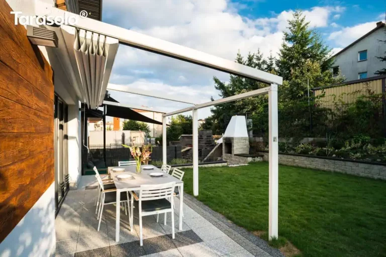 Biała pergola tarasowa Tarasola Simple o konstrukcji aluminiowej z całkowicie otwartym dachem, zamontowana do ściany budynku wykończonej panelami o strukturze drewna. Tkanina zadaszenia jest zwinięta w estetyczne fałdy przy elewacji, odsłaniając widok na niebo i aluminiowe szyny prowadzące. Konstrukcja opiera się na dwóch białych słupach nośnych, pod którymi na nowoczesnym tarasie z płyt ustawiono prostokątny stół jadalniany i sześć białych krzeseł. W tle widoczny jest zielony trawnik, murowany grill ogrodowy, kamienne ogrodzenie oraz wysokie drzewa iglaste pod lekko zachmurzonym niebem.