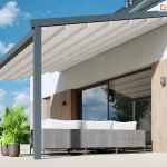 Wizualizacja prezentująca produkt pergola przyścienna Moderno zainstalowaną na nowoczesnym tarasie, ukazana pod kątem z perspektywy bocznej. Konstrukcja pergoli w kolorze ciemnego antracytu jest zamocowana do białej ściany budynku, która została ozdobiona pionowymi panelami o teksturze jasnego drewna. Zadaszenie stanowi jasna, kremowa tkanina ułożona w estetyczne fałdy, rozpięta na poprzecznych profilach, w których zamontowano zintegrowane punkty oświetlenia LED. Całość konstrukcji wspiera się na trzech solidnych, kwadratowych słupach nośnych, ustawionych na jasnym, gładkim podłożu tarasu. Pod pergolą znajduje się długa, szara sofa technorattanowa z białymi poduszkami oparciowymi, ustawiona tyłem do dużego przeszklenia drzwi tarasowych, w których odbija się błękitne niebo i okoliczna zieleń. Po lewej stronie, obok konstrukcji, stoją dwie brązowe donice z bujnymi zielonymi roślinami, w tym palmą. W tle widoczny jest fragment dachu sąsiedniego budynku oraz zielony żywopłot, a nad całością roztacza się pogodne niebo z lekkimi chmurami.