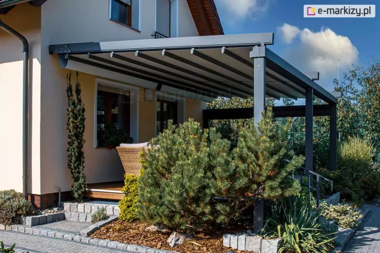 Zewnętrzna konstrukcja typu pergola z systemem zadaszenia materiałowego, zamontowana przy ścianie budynku jednorodzinnego w jasnym kolorze elewacji. Solidny stelaż pergoli wykonany jest z elementów aluminiowych malowanych proszkowo na kolor ciemnoszary lub antracytowy, co zapewnia nowoczesny i estetyczny wygląd całej instalacji. System dachowy stanowi rozsuwana tkanina o technicznej fakturze, która w widocznej części jest częściowo rozsunięta, tworząc system równoległych fałd wzdłuż prowadnic konstrukcyjnych. Pergola Solid Aluprof opiera się na trzech pionowych słupach nośnych, które są osadzone w otoczeniu ogrodu, bezpośrednio przy utwardzonej ścieżce wyłożonej kostką brukową. W bezpośrednim sąsiedztwie konstrukcji znajduje się bujna roślinność, w tym niewielkie drzewka iglaste, krzewy oraz ozdobne trawy, które naturalnie komponują się z architekturą domu. Widoczna część tarasu, osłonięta przez system zadaszenia, wykończona jest drewnianymi deskami tarasowymi, a w cieniu pergoli widać fragment wiklinowego fotela, co sugeruje zaaranżowanie przestrzeni wypoczynkowej. Oświetlenie dzienne na fotografii wskazuje na słoneczny dzień, podkreślając głębię kolorów konstrukcji oraz kontrast między ciemnym aluminium a jasną elewacją budynku, przy czym całość instalacji została zaprojektowana w celu zapewnienia ochrony przed nadmiernym nasłonecznieniem oraz poprawy komfortu użytkowania przydomowego tarasu.