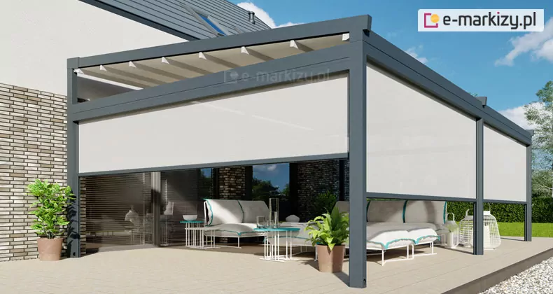Zdjęcie prezentuje produkt pergola tarasowa wolnostojąca Moderno w wersji dwumodułowej, zamontowaną jako zadaszenie tarasu przy nowoczesnym budynku. Konstrukcja o grafitowym kolorze wykonana jest z aluminium i wyposażona w systemowe rolety boczne typu screen w kolorze jasnoszarym, które są częściowo rozwinięte na wszystkich trzech widocznych bokach, tworząc kameralną przestrzeń. Zadaszenie pergoli stanowi biała tkanina ułożona w charakterystyczne fale, zamontowana na prowadnicach pod lekkim skosie w stronę frontu. Pod pergolą, na jasnym tarasie, znajduje się zestaw mebli wypoczynkowych z białymi poduszkami i błękitnymi detalami, a także dekoracyjne stoliki kawowe, donice z roślinami oraz ażurowe lampiony. Budynek, do którego przylega konstrukcja, posiada elewację z jasnej, wąskiej cegły oraz duże przeszklenia, a jego dach pokryty jest ciemną dachówką z oknem połaciowym. W tle widoczny jest zielony ogród z gęstym żywopłotem i drzewami pod błękitnym niebem z nielicznymi chmurami.