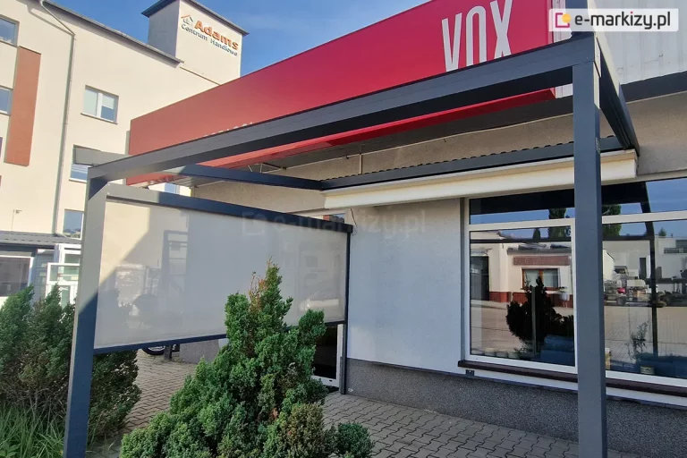 Zdjęcie prezentuje zewnętrzną konstrukcję pergoli tarasowej Square zamontowanej przy elewacji budynku handlowego, wykonaną z profili aluminiowych w kolorze ciemnego grafitu. Pergola posiada płaski, zwinięty do ściany dach oraz dodatkową pionową osłonę boczną typu screen w kolorze jasnoszarym, która jest częściowo rozwinięta, zapewniając ochronę przed słońcem i wiatrem. Konstrukcja jest przymocowana do jasnej, tynkowanej ściany budynku, na której powyżej widnieje duży czerwony kaseton reklamowy z białym napisem VOX. Pod pergolą znajduje się duże okno wystawowe w białej ramie, w którym odbija się otoczenie parkingu i sąsiednie obiekty. Podłoże pod konstrukcją wyłożone jest szarą kostką brukową, a bezpośrednio przed jednym ze słupów nośnych rośnie gęsty, zielony krzew iglasty. W tle, po lewej stronie, widoczny jest wielokondygnacyjny biały budynek z napisem Adams Centrum Handlowe na elewacji. Scena została uchwycona w pełnym świetle dziennym przy bezchmurnym, błękitnym niebie, co podkreśla techniczne detale wykończenia systemów osłonowych oraz ich integrację z architekturą obiektu.