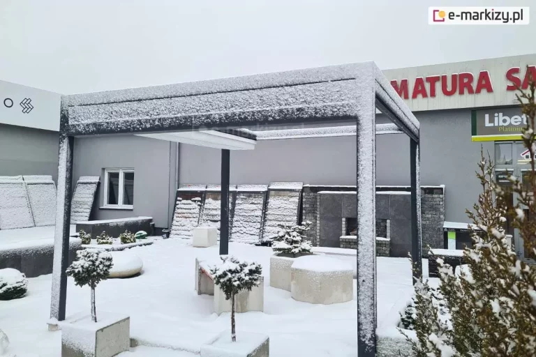 Zdjęcie prezentuje nowoczesną pergolę tarasową Luxury w warunkach zimowych, pokrytą warstwą śniegu, co potwierdza jej całoroczną wytrzymałość konstrukcyjną. Wolnostojąca rama wykonana z profili aluminiowych w kolorze antracytowym jest oprószona białym puchem, podobnie jak otaczający ją teren i elementy ekspozycji ogrodowej. Zadaszenie tkaninowe pergoli jest całkowicie zwinięte i ukryte pod systemowym daszkiem ochronnym, co zabezpiecza mechanizm i materiał przed ciężarem zalegającego śniegu oraz wilgocią w okresie zimowym. Konstrukcja stoi na ośnieżonym placu obok nowoczesnego budynku, a w jej bliskim sąsiedztwie widoczne są betonowe donice z krzewami oraz ekspozycyjne płyty elewacyjne, również pokryte śniegiem. Całość obrazu podkreśla stabilność i trwałość konstrukcji Luxury w trudnych warunkach atmosferycznych.