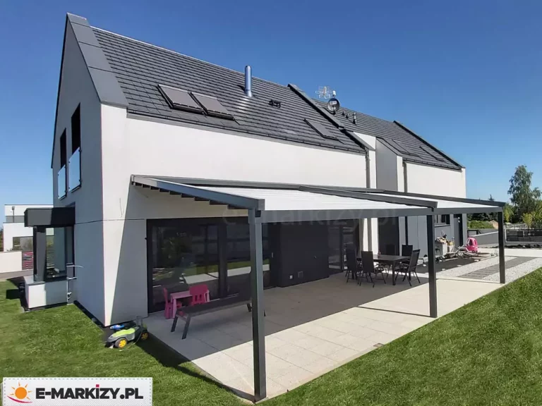Szeroka, dwumodułowa pergola tarasowa o antracytowej, aluminiowej konstrukcji, zamontowana do elewacji nowoczesnego domu piętrowego z dwuspadowym dachem pokrytym ciemną dachówką. Zadaszenie pergoli wykonane jest z białego materiału, który jest całkowicie rozwinięty i rozpięty na poziomych profilach, tworząc dużą strefę cienia nad tarasem. Konstrukcja wspiera się na trzech masywnych, pionowych słupach. Pod pergolą, na tarasie wyłożonym dużymi, jasnymi płytami betonowymi, znajduje się strefa jadalniana z ciemnym stołem i sześcioma krzesłami oraz kącik dziecięcy z małym różowym stolikiem i krzesełkami. Na trawniku obok tarasu widoczna jest dziecięca zabawka jeździk w kolorze zielono-czarnym. Elewacja budynku jest biała, z ciemnoszarymi wnękami okiennymi i dużymi przeszkleniami na parterze, w których odbija się ogród. Na dachu domu zamontowane są kolektory słoneczne oraz antena satelitarna. Całość ujęcia została wykonana z góry, z perspektywy lotu ptaka, w pełnym słońcu na tle bezchmurnego nieba.