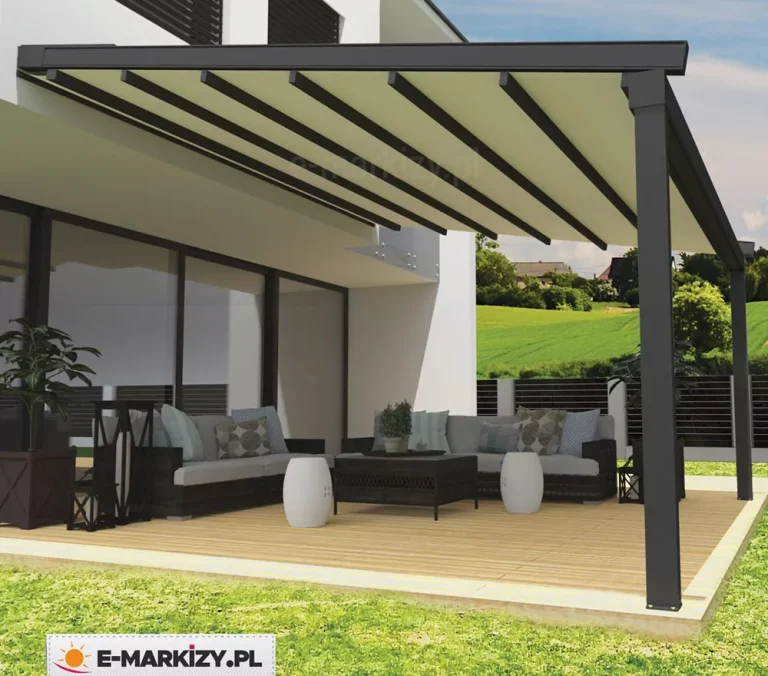 Nowoczesna pergola tarasowa Square o konstrukcji aluminiowej w kolorze antracytowym, zamontowana do elewacji białego budynku o minimalistycznej bryle. Zadaszenie pergoli składa się z jasnego, kremowego poszycia materiałowego rozpiętego na systemie równoległych, ciemnych profili poprzecznych, które nadają konstrukcji rytmiczny wygląd. Pod zadaszeniem zaaranżowano przestronną strefę wypoczynkową na tarasie wyłożonym jasnymi deskami kompozytowymi. W skład wyposażenia wchodzi duży, narożny zestaw mebli ogrodowych z ciemnego technorattanu z szarymi poduszkami siedziskowymi i dekoracyjnymi poduszkami w geometryczne wzory. Przed kanapą ustawiono niski, prostokątny stolik kawowy oraz dwa białe, ceramiczne pufy w kształcie bębnów. Po lewej stronie widoczna jest duża, ciemna donica z rośliną o szerokich liściach oraz dekoracyjne latarnie ogrodowe. W tle rozpościera się widok na zielone wzgórza, łąki oraz pojedynczą zabudowę wiejską pod błękitnym niebem z delikatnymi chmurami. Teren posesji oddzielony jest od otoczenia nowoczesnym, ciemnym ogrodzeniem panelowym, a bezpośrednio przy tarasie znajduje się pas zielonego, zadbanego trawnika.