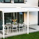 Pergola Simple Tarasola Galeria Zdjęć: Szeroka, biała pergola tarasowa Tarasola Simple o konstrukcji przyściennej, zamontowana na elewacji nowoczesnego domu jednorodzinnego. Zadaszenie składa się z dwóch sekcji białego, napiętego materiału technicznego, zamocowanego na aluminiowych profilach poprzecznych i prowadnicach. Konstrukcja opiera się na trzech pionowych słupach o kwadratowym przekroju, które są zakotwiczone w gruncie, na trawniku przy krawędzi tarasu. Pod zadaszeniem znajduje się strefa wypoczynkowa z dużym, prostokątnym stołem o szarym blacie oraz sześcioma białymi krzesłami z ciemnymi poduszkami. Taras został wyłożony jasnymi, nakrapianymi płytami z ozdobnymi ciemniejszymi pasami. Elewacja budynku łączy biały tynk z panelami imitującymi naturalne drewno w ciepłym brązowym odcieniu. W tle widoczne są duże przeszklenia drzwi tarasowych, fragment zielonego trawnika oraz sąsiednia zabudowa. Zdjęcie wykonane z perspektywy bocznej, ukazujące znaczną szerokość konstrukcji i jej integrację z bryłą budynku.