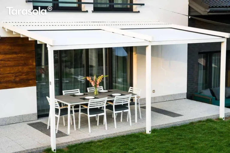 Pergola Simple Tarasola Galeria Zdjęć: Szeroka, biała pergola tarasowa Tarasola Simple o konstrukcji przyściennej, zamontowana na elewacji nowoczesnego domu jednorodzinnego. Zadaszenie składa się z dwóch sekcji białego, napiętego materiału technicznego, zamocowanego na aluminiowych profilach poprzecznych i prowadnicach. Konstrukcja opiera się na trzech pionowych słupach o kwadratowym przekroju, które są zakotwiczone w gruncie, na trawniku przy krawędzi tarasu. Pod zadaszeniem znajduje się strefa wypoczynkowa z dużym, prostokątnym stołem o szarym blacie oraz sześcioma białymi krzesłami z ciemnymi poduszkami. Taras został wyłożony jasnymi, nakrapianymi płytami z ozdobnymi ciemniejszymi pasami. Elewacja budynku łączy biały tynk z panelami imitującymi naturalne drewno w ciepłym brązowym odcieniu. W tle widoczne są duże przeszklenia drzwi tarasowych, fragment zielonego trawnika oraz sąsiednia zabudowa. Zdjęcie wykonane z perspektywy bocznej, ukazujące znaczną szerokość konstrukcji i jej integrację z bryłą budynku.