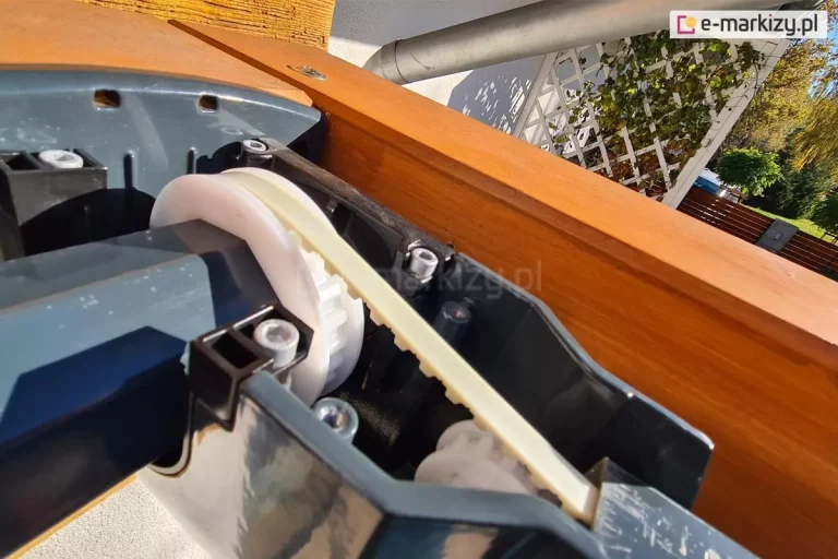 Zbliżenie mechanizmu paskowego ukrytego w prowadnicach dachu do pergoli Silva. Prowadnica zamocowana do drewnianej pergoli na tarasie z widocznym w tle ogrodem.