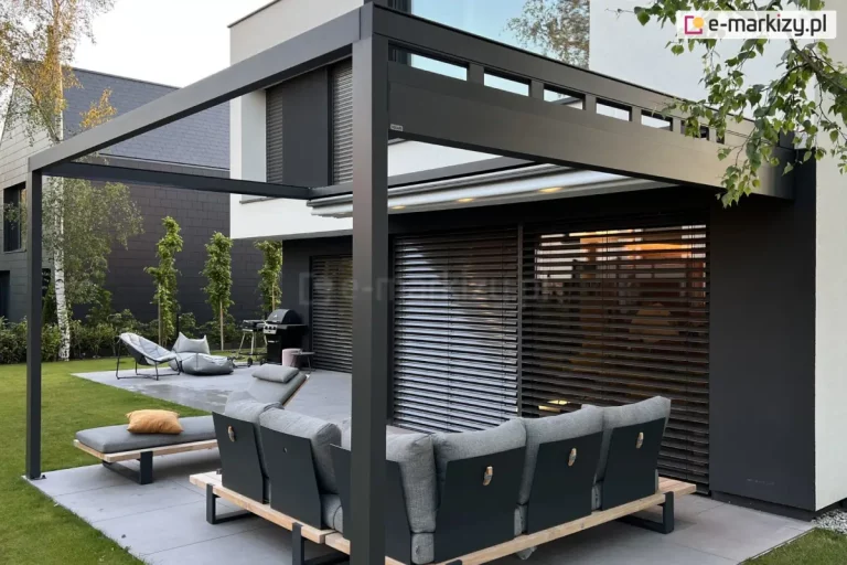 Nowoczesny taras przylegający do nowoczesnego domu w minimalistycznym stylu. Konstrukcja zadaszenia to ciemnoszara, aluminiowa pergola z prostokątnym, otwartym dachem i poziomymi belkami u góry. Główne słupy podtrzymujące pergolę są masywne i proste, utrzymane w tej samej kolorystyce. Pod zadaszeniem ustawiono dużą sofę modułową w kształcie litery L z drewnianą podstawą, ciemnymi bokami i jasnoszarymi poduszkami – jeden z segmentów służy jako leżanka, na której leży żółta poduszka dekoracyjna. W tle za sofą znajduje się elewacja domu z dużym, przeszklonym przesuwanym oknem, zasłoniętym poziomymi, zewnętrznymi żaluzjami w kolorze ciemnego brązu. Po lewej stronie zdjęcia widoczne są dwa nowoczesne siedziska na otwartym tarasie, ustawione na dużych, szarych płytach. Obok nich znajduje się grill gazowy oraz ozdobna donica. W tle – pas zieleni z kolumnowymi iglakami i brzozami na tle grafitowego ogrodzenia. Przestrzeń zaprojektowana jest w duchu nowoczesnego minimalizmu, z naciskiem na funkcjonalność i estetykę.