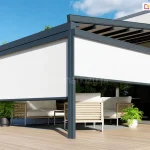 Nowoczesna pergola tarasowa Square o antracytowej, wykonanej z aluminium konstrukcji, wyposażona w białe przesłony boczne typu screen. Przesłony są częściowo opuszczone, zasłaniając górną połowę prześwitów między słupami i tworząc intymną strefę cienia. Konstrukcja zadaszenia posiada widoczne poprzeczne belki w kolorze ciemnoszarym, a całość jest zamontowana do elewacji nowoczesnego budynku o jasnoszarym i grafitowym wykończeniu. Pod pergolą znajduje się luksusowy zestaw mebli ogrodowych z jasnymi poduszkami i drewnianymi elementami wykończeniowymi na podłokietnikach. Na jasnym, drewnianym tarasie ustawione są również donice z zielonymi roślinami – jedna ciemna po lewej stronie i biała po prawej. W tle widoczny jest gęsty zielony żywopłot oraz fragment basenu w prawym dolnym rogu zdjęcia. Scena przedstawiona jest w pełnym słońcu na tle czystego, błękitnego nieba.