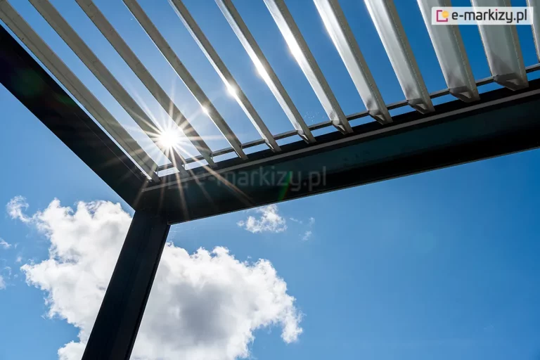 Pergola tarasowa SB 400 Aluprof z antracytową konstrukcja oraz otwartym dachem panelowym z białych lameli, przepuszczającym promienie słoneczne na tle błękitnego nieba z pierzastymi białymi chmurkami.