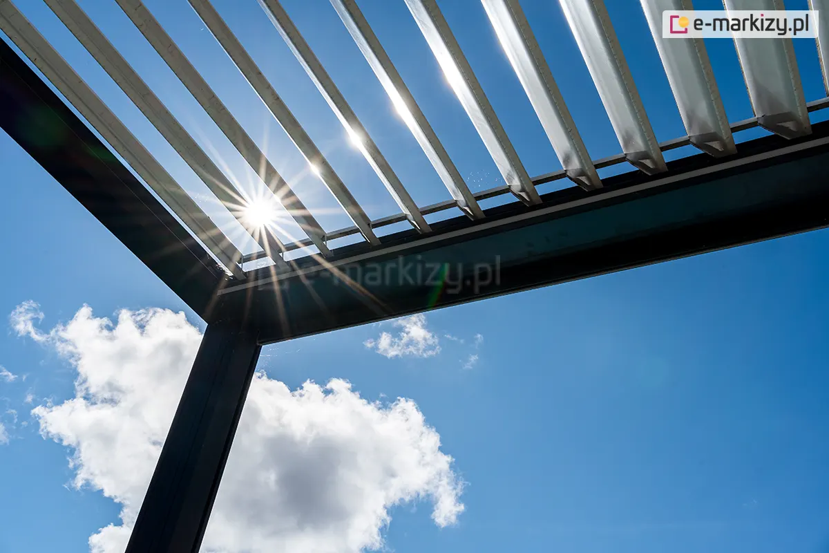 Pergola tarasowa SB 400 Aluprof z antracytową konstrukcja oraz otwartym dachem panelowym z białych lameli, przepuszczającym promienie słoneczne na tle błękitnego nieba z pierzastymi białymi chmurkami.