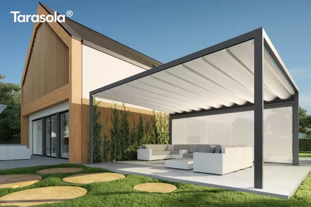 Pergola tarasowa wolnostojąca Tarasola Flat, zainstalowana na przestronnym tarasie z jasnych, kompozytowych desek. Aluminiowa rama w kolorze głębokiego antracytu wspiera białe, tkaninowe zadaszenie rzymskie, które jest rozłożone i układa się w charakterystyczne, miękkie fale podwieszone na poprzecznych profilach. Konstrukcja charakteryzuje się masywną górną belką, która pełni funkcję estetyczną oraz techniczną, skrywając system odprowadzania wody. Pod pergolą zaaranżowano luksusową strefę wypoczynku z białym meblem narożnym i niskim stolikiem kawowym, osłoniętą w pełni rozwiniętym refleksolem Zip. W tle widoczny jest nowoczesny budynek z elewacją łączącą biel, cegłę oraz duże przeszklenia, a także starannie zaprojektowany ogród z trawnikiem i ozdobnymi trawami.